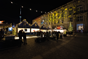 Ein lebendiger Weihnachtsmarkt in Berlin, Deutschland, mit Menschen um beleuchtete Stände, festlicher Dekoration und Gebäuden mit Fenstern unter einem dunklen Himmel.