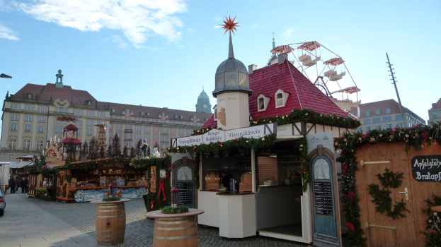 Ein belebter Weihnachtsmarkt auf einem Stadtplatz mit geschmückten Bäumen, Gebäuden, Straßenlaternen, Fahrzeugen, Passanten und einem bewölkten Himmel.