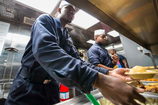 Eine Gruppe Von M├Ąnnern Steht In Einer K├╝che, Einer Hält Ein Teller Mit Essen, Mit Tabletts Mit Essen Auf Dem Tisch, Einem Feuerl├Âscher Und Papieren An Der Wand Im Hintergrund.