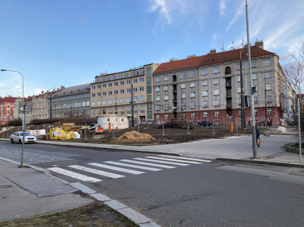 Baustelle für ein neues Wohnhaus in einer Stadtstraße mit parkenden Autos, Laternen, Bäumen, Gebäuden und einem bewölktem Himmel.