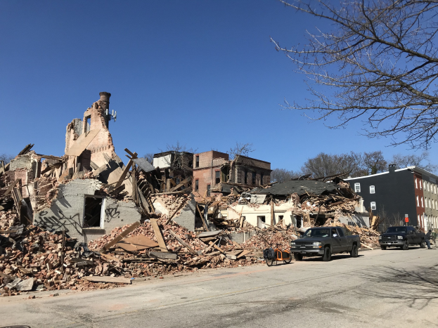 Ein eingestürztes Gebäude an einer Straßenecke mit Fahrzeugen, Fußgängern, Bäumen und einem klaren Himmel, umgeben von Schutt und Trümmern.