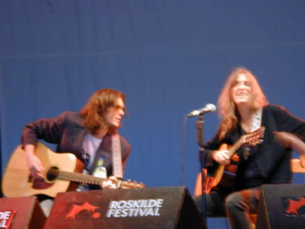 Zwei Personen auf der Bühne mit musikalischen Instrumenten, einem Mikrofon und Kisten mit der Aufschrift 'ROCK SLIDE FESTIVAL'.