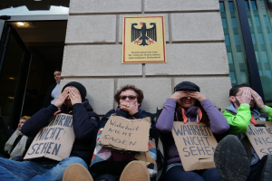 Eine Gruppe von Menschen sitzt vor einem Gebäude, hält Schilder, einige bedecken ihre Gesichter, mit einem Logo und einer Tafel an der Wand und einer Glastür auf der rechten Seite.