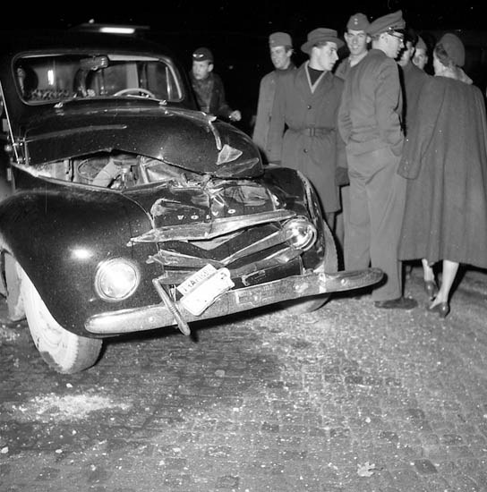 Schwarzes und weißes Bild eines schwer beschädigten Autos am Straßenrand mit eingedellter Motorhaube und zersplitterten Fenstern, umgeben von einer Gruppe von Menschen, die die Szene begutachten.