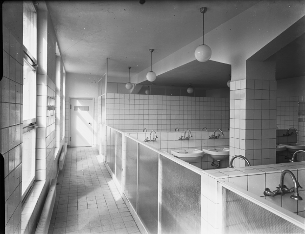 Ein Schwarz-Weiß-Foto einer öffentlichen Toilette mit Waschbecken, Wasserhähnen, von der Decke hängenden Lampen, Wänden, Fenstern und einer Tür.