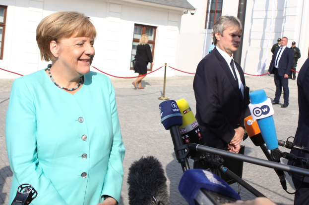 Bundeskanzlerin Angela Merkel spricht vor Reportern im Freien, umgeben von einer Gruppe mit Mikrofonen und einer Absperrung, unter einem bewölkten Himmel.