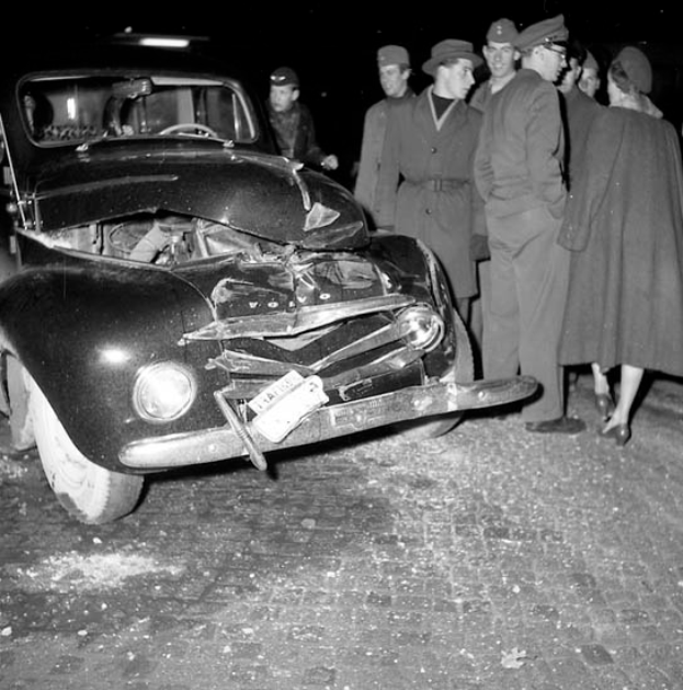 Ein Schwarz-Weiß-Bild eines verbeulten Autos am Straßenrand mit zersplitterten Fenstern und einer Gruppe besorgter Menschen drumherum.