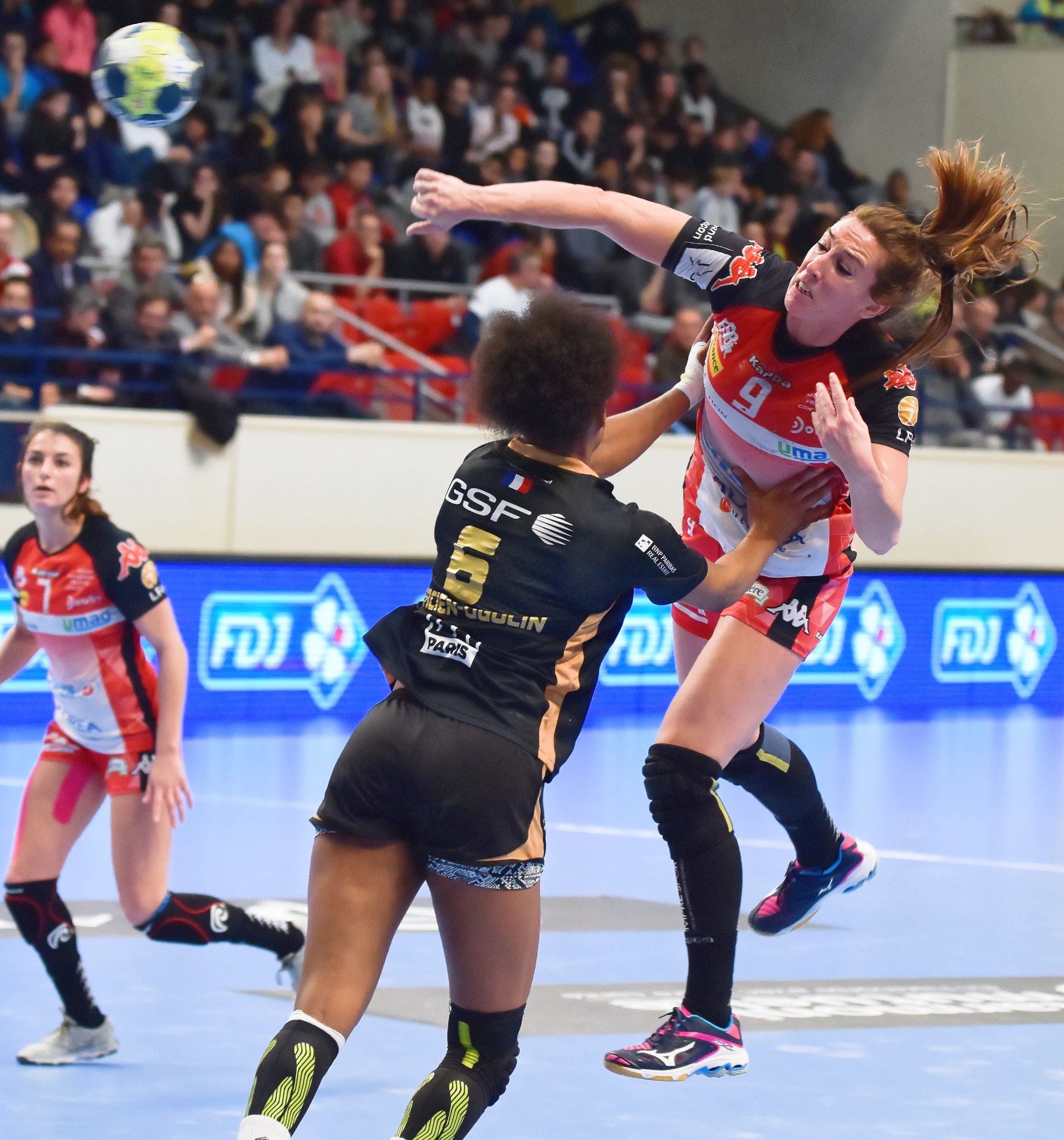 Frauen beim Handballspielen auf einem Feld mit einem Ball in der Luft, in einer Arena mit Zuschauern auf Stühlen, Geländern und einer Wand.
