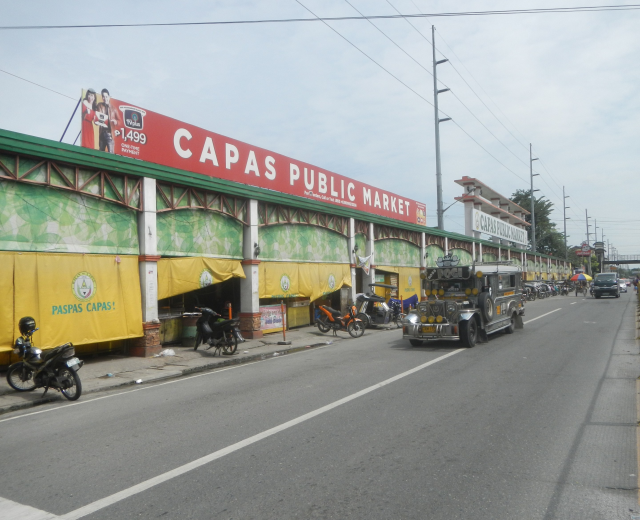 Eine belebte Stadtstraße mit Fahrzeugen, einem Gehweg, Strommasten, Gebäuden, Bäumen und einem bewölkten Himmel, mit einem Gebäude mit der Aufschrift "Capas Public Market" im Vordergrund.