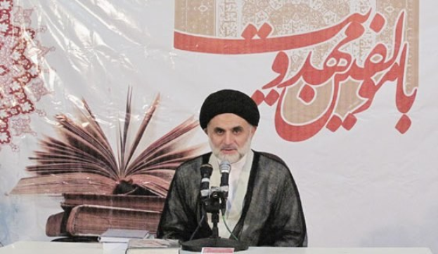 Ayatollah Ali Khamenei, Oberster Führer von Iran, sitzt vor einem Tisch mit Büchern und Mikrofonen, mit einem Banner mit Text und einem Buchbild dahinter.