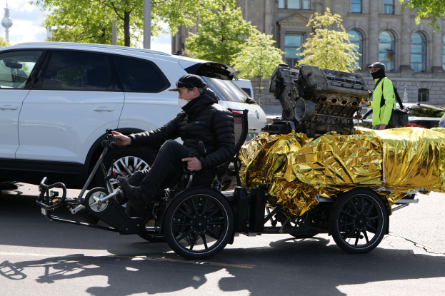 Ein Mann in einer schwarzen Jacke und Mütze sitzt in einem Rollstuhl mit einem großen Motor an der Rückseite, umgeben von Fahrzeugen auf einer Straße mit Bäumen, Gebäuden und Polen im Hintergrund unter einem klaren blauen Himmel.