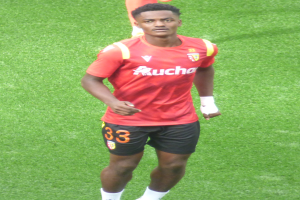 Emmanuel Adebayor, ein Mann in einem roten Hemd und schwarzen Shorts, geht auf einem Fussballfeld mit zwei Personen im Hintergrund.