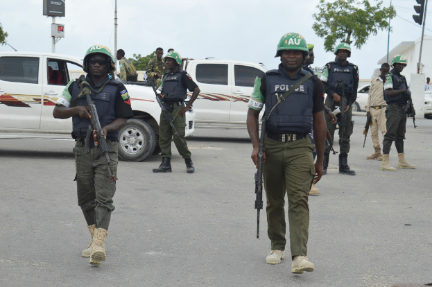Eine Gruppe von Polizisten in Helmen, die Waffen halten, geht eine Straße entlang mit Fahrzeugen, Laternenmästen, Ampeln, Texttafeln, Bäumen, Gebäuden und einem bewölkten Himmel im Hintergrund.