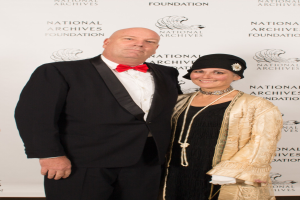 Ein Mann in einem schwarzen Anzug mit weißem Hemd und schwarzer Fliege steht neben einer Frau in einem eleganten Kleid mit einem gemusterten Schal auf einem roten Teppich, mit einem weißen Banner mit Text im Hintergrund.