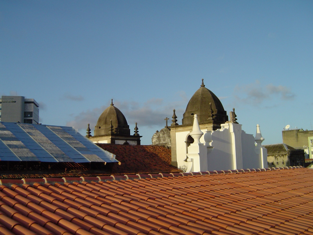 Stadtansicht mit Gebäuden im Vordergrund und einem blauen Himmel im Hintergrund, mit Solarpanelen auf einem Dach, die den Einsatz von erneuerbarer Energie anzeigen.