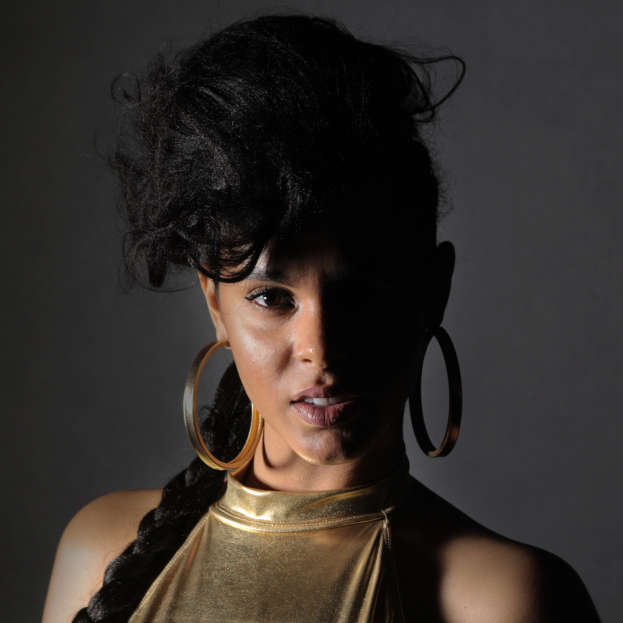 Eine Frau in einem goldenen Kleid und großen Ohrringen steht selbstbewusst vor einer Wand, ihr Haar ist zu einem Bob gestylt und sie schaut direkt in die Kamera mit einem entschlossenen Gesichtsausdruck.