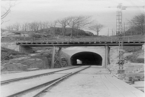 Ein Schwarz-Weiß-Foto eines Tunnels im Bau mit Eisenbahnschienen, einer Brücke mit Säulen und Geländern, Bäumen im Hintergrund und einem Himmel darüber.