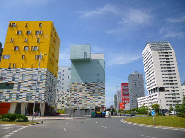 Eine Stadtstraße mit hohen Gebäuden im Hintergrund, Straßenlaternen, Autos, Informationsschildern, Büschen, Bäumen und einem bewölkten Himmel, mit einzigartig gestalteten Gebäuden der Universität Rotterdam.