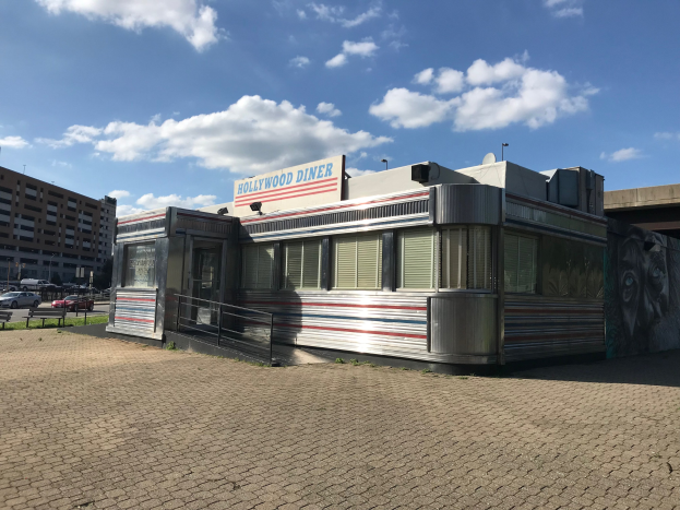 Außenansicht des Hollywood Diners mit seiner Leuchtschrift, umgeben von Gebäuden, Straßenmöbeln, Fahrzeugen und einem bewölkten Himmel.