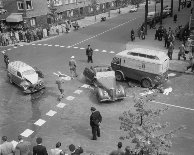 Eine Schwarz-Weiß-Szene eines Autounfalls an einer Straßenkreuzung, mit mehreren Fahrzeugen einschließlich eines Vans, einer Gruppe von Menschen drumherum und im Hintergrund Gebäuden, Bäumen, Pfählen und Texttafeln.