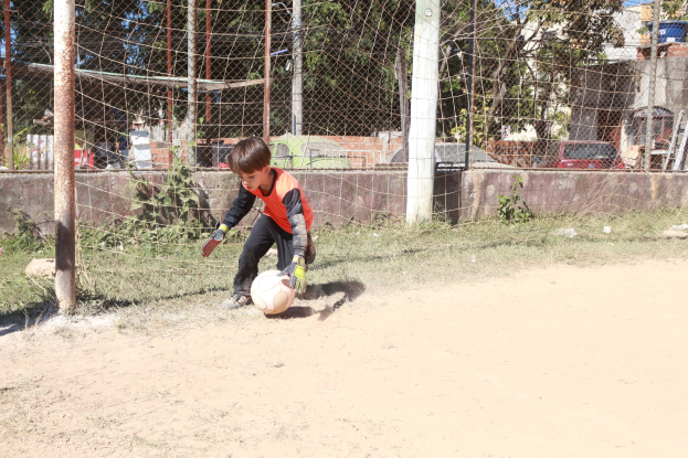 Junger Junge beim Fußballspielen auf einem Schmutzfeld mit Gras, Pflanzen, Pfählen, einem Zaun, einer Wand, Bäumen, Fahrzeugen, Gebäuden und einer bewölkten Himmel