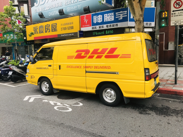 Gelber DHL-Lieferwagen auf der Straße geparkt, umgeben von Gebäuden, Bäumen, Schildern, Strommasten, Ampeln und einem Fußgänger auf dem Gehweg.