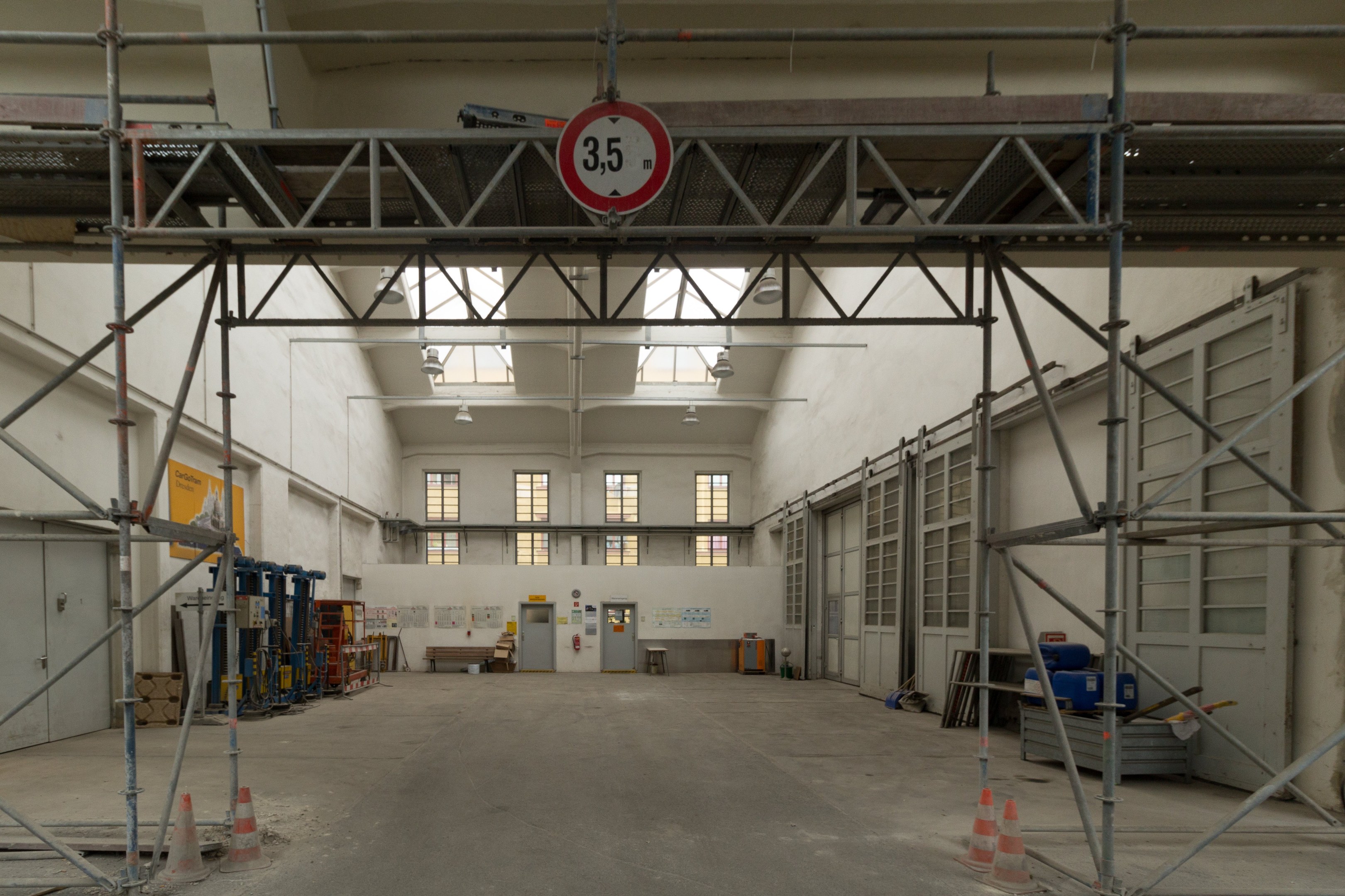 Großes Lagerinneres mit ausgedehnten Gerüsten, Verkehrskegeln, Metallstangen, einem Schild, Maschinen, einer Tür, Fenstern, Wänden, Deckenleuchten und einem Dach.