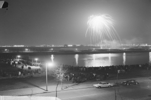 Ein Schwarz-Weiß-Foto einer Menge, die ein Feuerwerk über Wasser beobachtet, mit Fahrzeugen, Laternenpfählen, Bäumen und Gebäuden im Hintergrund.