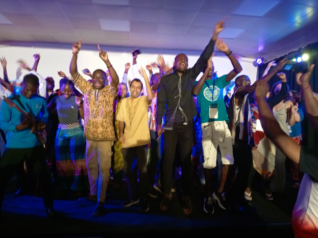 Eine Gruppe von Menschen, die auf einer Bühne tanzen und die Hände heben, beleuchtet von Scheinwerfern auf der rechten Seite und einem Bildschirm im Hintergrund, unter einer Decke mit Beleuchtung.