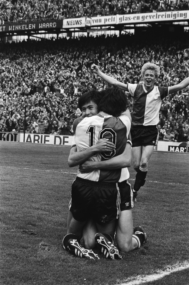 Drei Fussballspieler umarmen sich auf dem Boden und feiern ein Tor vor einer jubelnden Menge in einer schwarz-weissen Stadionszene.