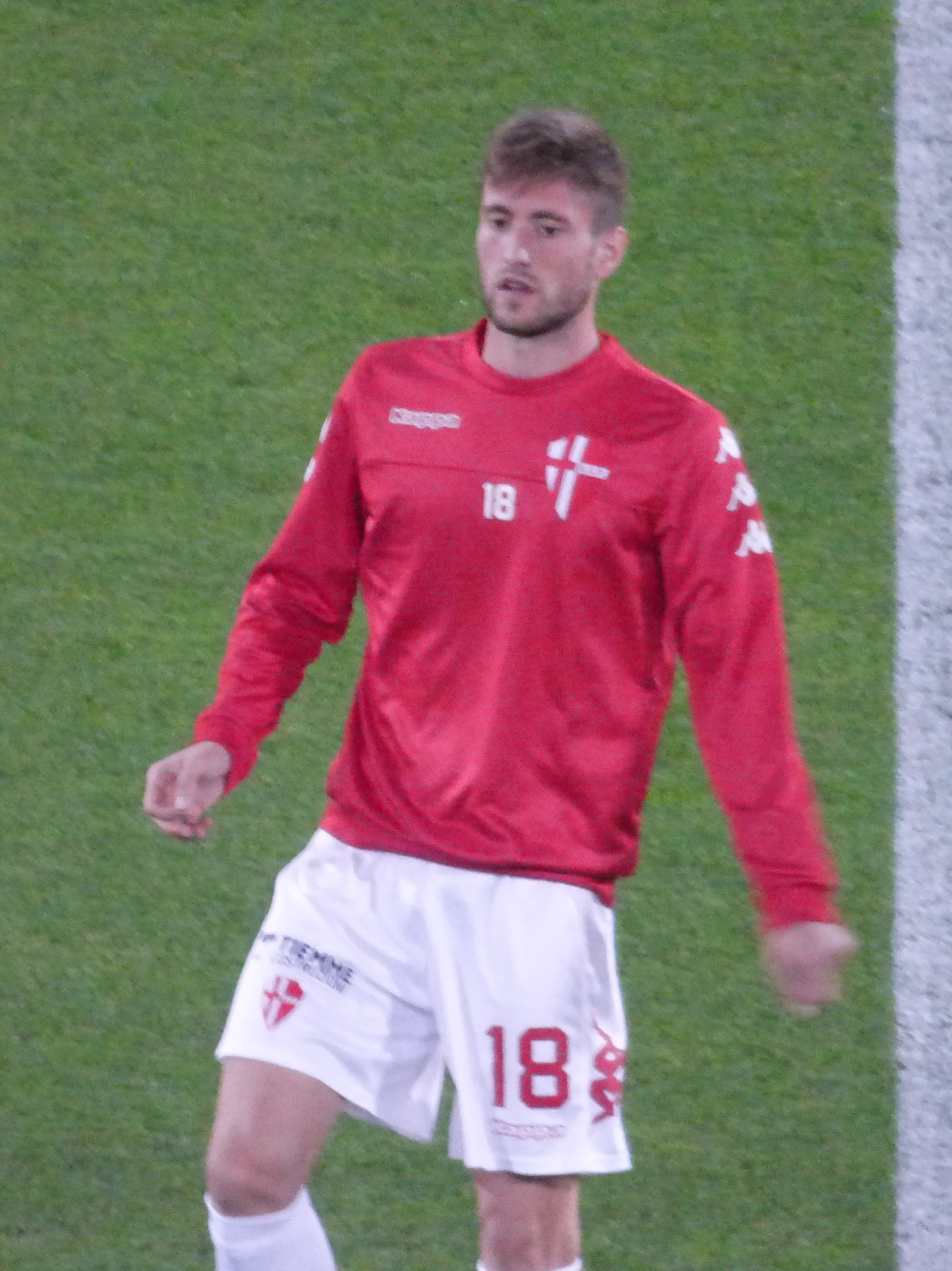 Ein Fußballspieler in rotem Trikot und weißen Shorts steht auf einem Feld mit ausgestreckten Armen und schaut nach vorne.