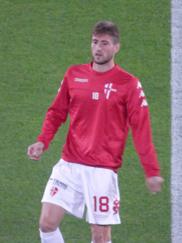 Ein Fußballspieler in rotem Trikot und weißen Shorts steht auf einem Feld mit ausgestreckten Armen und schaut nach vorne.