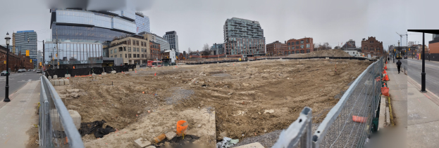 Baustelle mit einem großen Haufen Erde, umgeben von Gebäuden, Straßeninfrastruktur, Fahrzeugen, Passanten, Bäumen und einem klaren blauen Himmel.
