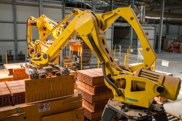 Großes Industriegebäude mit gestapelten Kisten, Kränen an der Decke, Zaun, Versorgungsmasten und einer fernen Wand in einer automatisierten Produktionsumgebung.