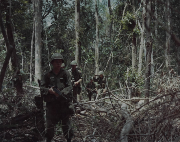Eine Gruppe von Soldaten in Helmen und mit Gewehren bewaffnet durchquert einen üppigen Wald mit hohen Bäumen, mit Text am unteren Bildrand.