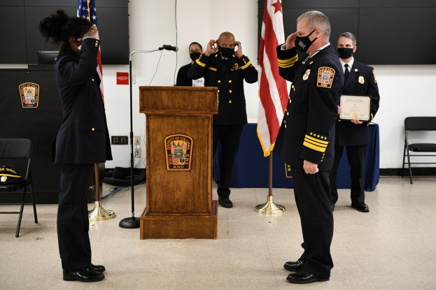 Eine Gruppe uniformierter und maskierter Personen steht vor einem Podium mit einem Mikrofon, umgeben von Flaggen, Stühlen und Tischen, während eine neue Feuerwache vereidigt wird, wobei eine Person ein Zertifikat hält.