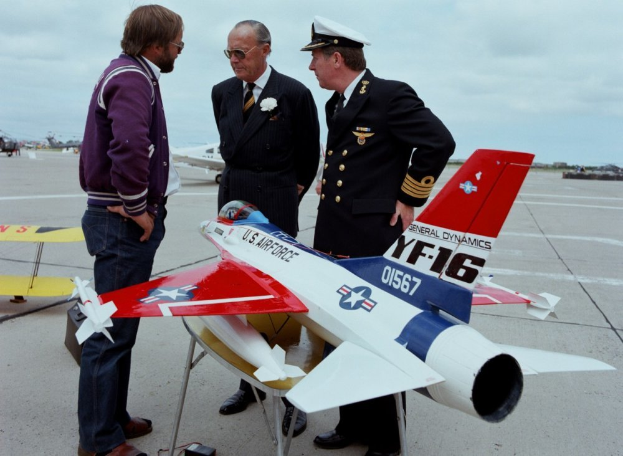 Drei Männer im Businessanzug stehen um ein Modellflugzeug auf einem Rollfeld, mit einem weißen und grauen Himmel im Hintergrund.