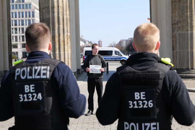 Drei Polizeibeamte stehen vor einem Gebäude, wobei ein Mann in der Mitte ein Papier hält, flankiert von Säulen, während Fahrzeuge, Menschen, Bäume und Gebäude den Hintergrund unter dem Himmel ausfüllen.
