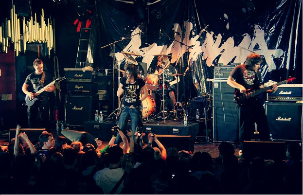 Eine Gruppe von Musikern auf der Bühne mit Gitarren und Trommeln, Mikrofonen und Ständern, mit einem Banner im Hintergrund und Text unten lesend "The Wanted - Live in der O2 Academy, London".