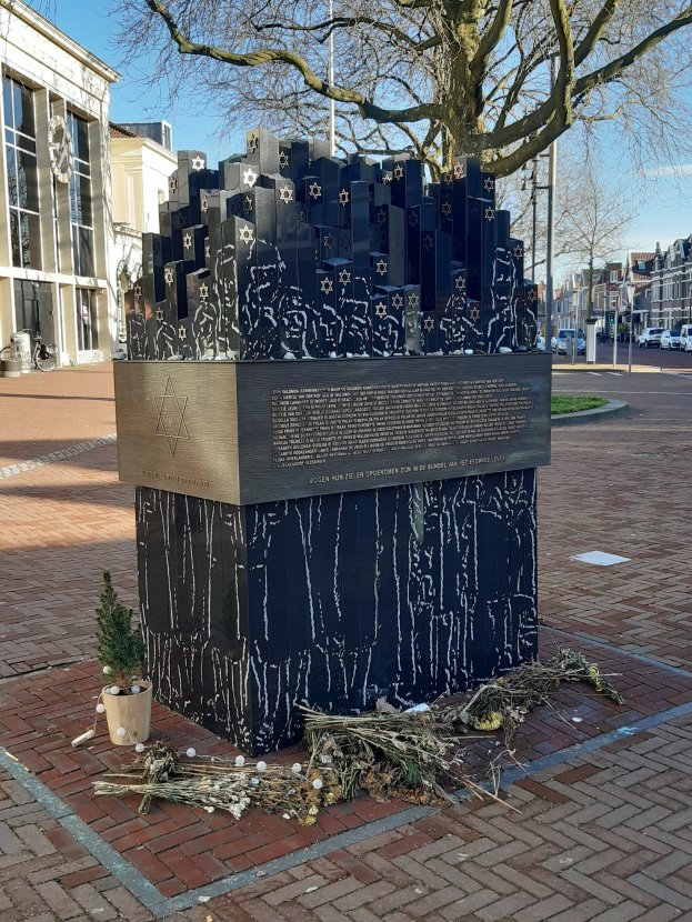 Denkmal in einem Stadtplatz, das an Holocaust-Opfer erinnert, mit einem Baum davor, einem Topfgewächs am Boden, verstreuten trockenen Blättern und Gebäuden, Fahrzeugen, Pfählen und Bäumen im Hintergrund.