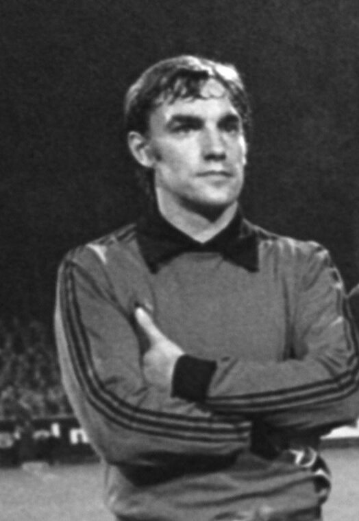 Schwarzes und weißes Foto eines Mannes in einem Anzug, der auf einem Fußballfeld mit verschränkten Armen steht und von einer Gruppe von Sitzenden im Hintergrund umgeben ist.