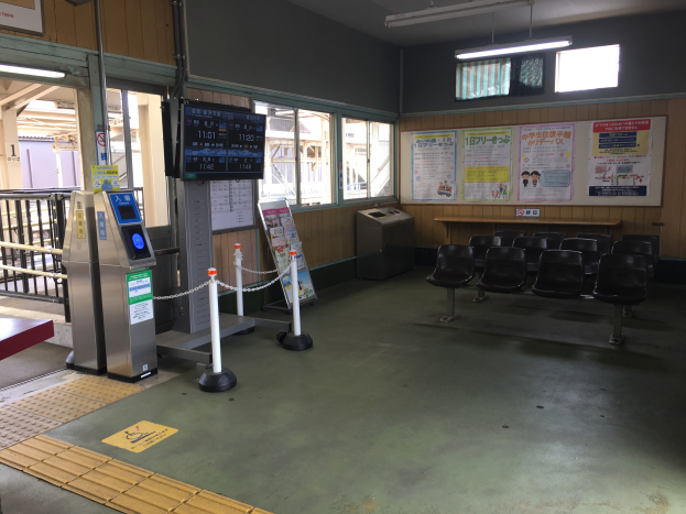 Ein belebter Bahnhofseingang mit Reihen von Stühlen, einer Informations counter, Ticketautomaten, Kettenpfählen, Informationsanzeigen, Wandplakaten, Deckenbeleuchtung, Geländern und anderen Einrichtungsgegenständen.
