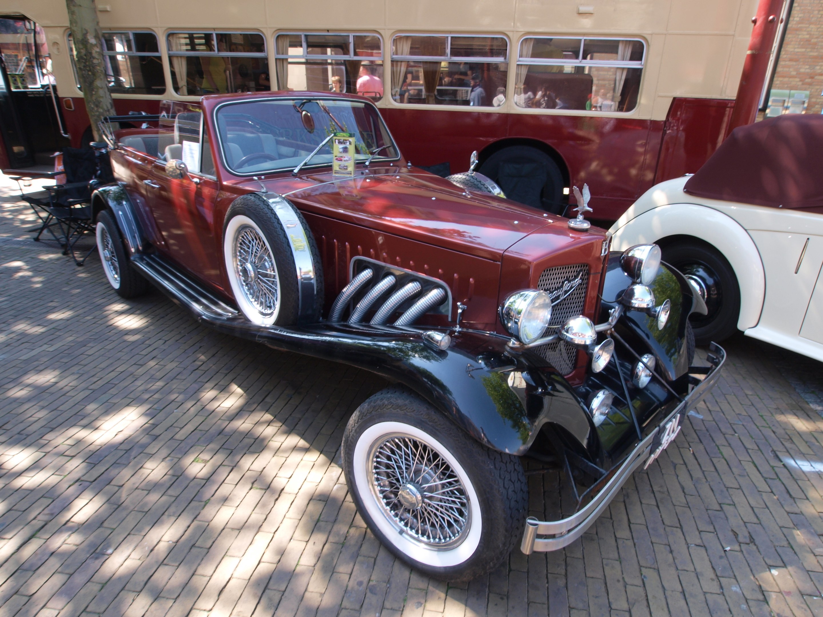 Ein Oldtimer neben einem Doppeldeckerbus auf einer Kopfsteinpflasterstraße, mit Stühlen und einem Baum im Vordergrund, einem Gebäude im Hintergrund und Menschen im Bus.
