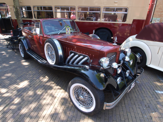 Ein Oldtimer neben einem Doppeldeckerbus auf einer Kopfsteinpflasterstraße, mit Stühlen und einem Baum im Vordergrund, einem Gebäude im Hintergrund und Menschen im Bus.