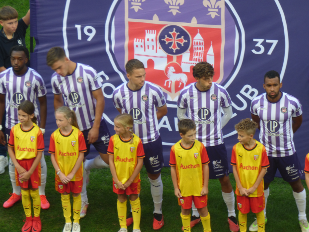 Eine Gruppe junger Menschen, die auf einem Fußballfeld zusammenstehen und ein Banner mit der Aufschrift "Preston Primary School Football Team" im Hintergrund haben.