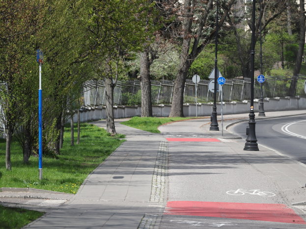 Stadtstraße mit einem Fahrradweg, der von Bäumen, Pfählen, Schildern, Gras, Pflanzen und einem Zaun gesäumt ist und zu Gebäuden unter einem klaren blauen Himmel führt.