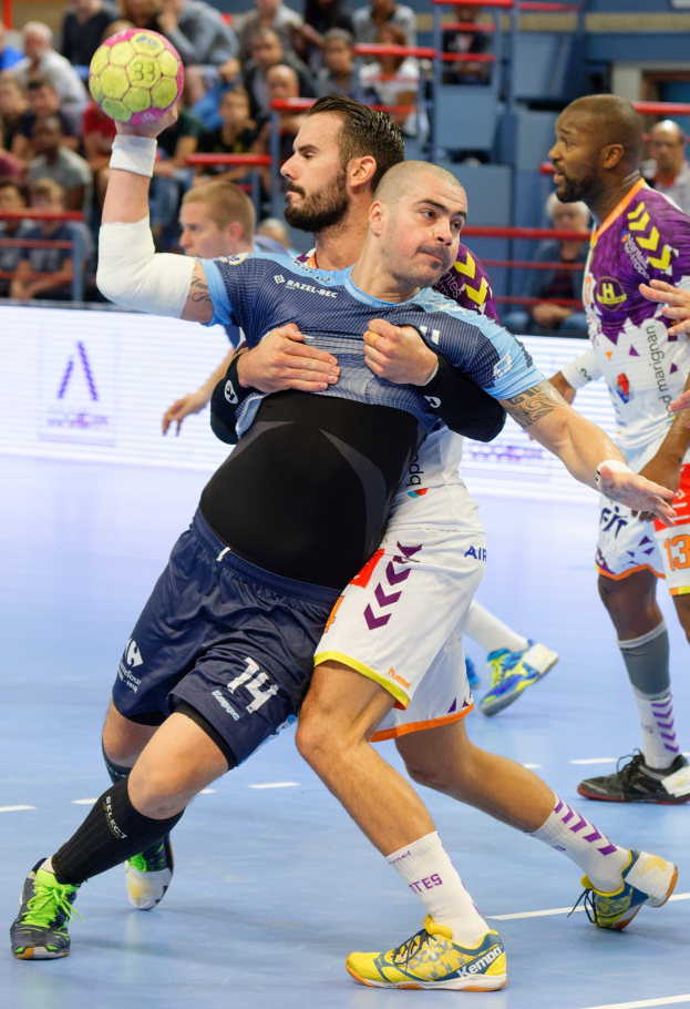 Eine Gruppe von Männern, die Handball in einem Stadion spielen, wobei ein Spieler den Ball hält.