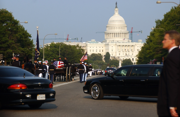 Trauerzug mit Menschen in Mützen und Fahnen vor dem Kapitolsgebäude, Fahrzeuge auf der Straße, Laternenpfähle, Bäume und Gebäude mit Fenstern unter einem klaren Himmel.