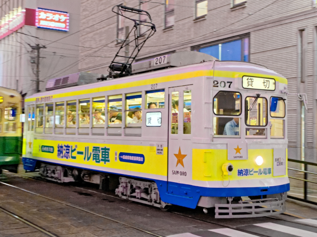 Eine gelbe und blaue Straßenbahn auf einer Stadtstraße bei Nacht mit Menschen darin, Gebäude mit Fenstern, Strommasten mit Drähten und Schilder im Hintergrund.
