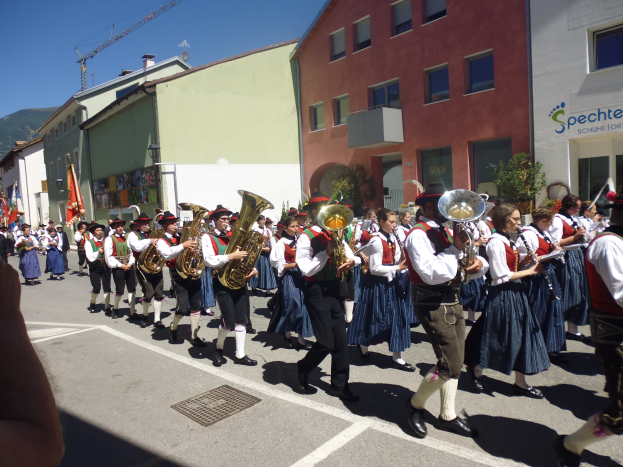 Eine Gruppe von Menschen in traditioneller bayrischer Tracht, die auf der Straße musizieren und dabei durch eine von Gebäuden gesäumte Straße gehen, einige halten Fahnen, im Hintergrund ein Hügel und ein klarer blauer Himmel.